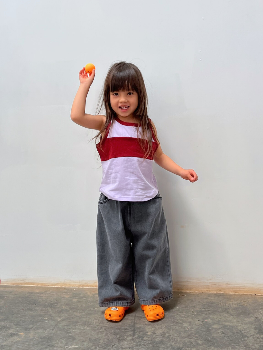 A young child stands indoors, proudly holding an orange ball while wearing PEPPER JEAN pants, a red and white shirt, and orange clogs.