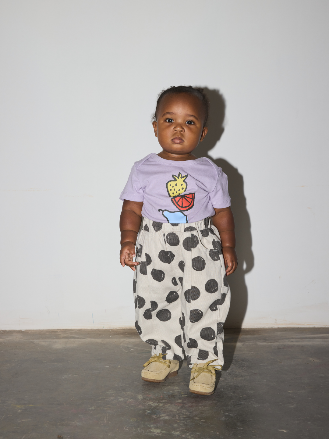 A toddler standing against a plain light gray wall wearing a light purple t-shirt with a fruit graphic and loose white pants with large black polka dots, paired with beige shoes.