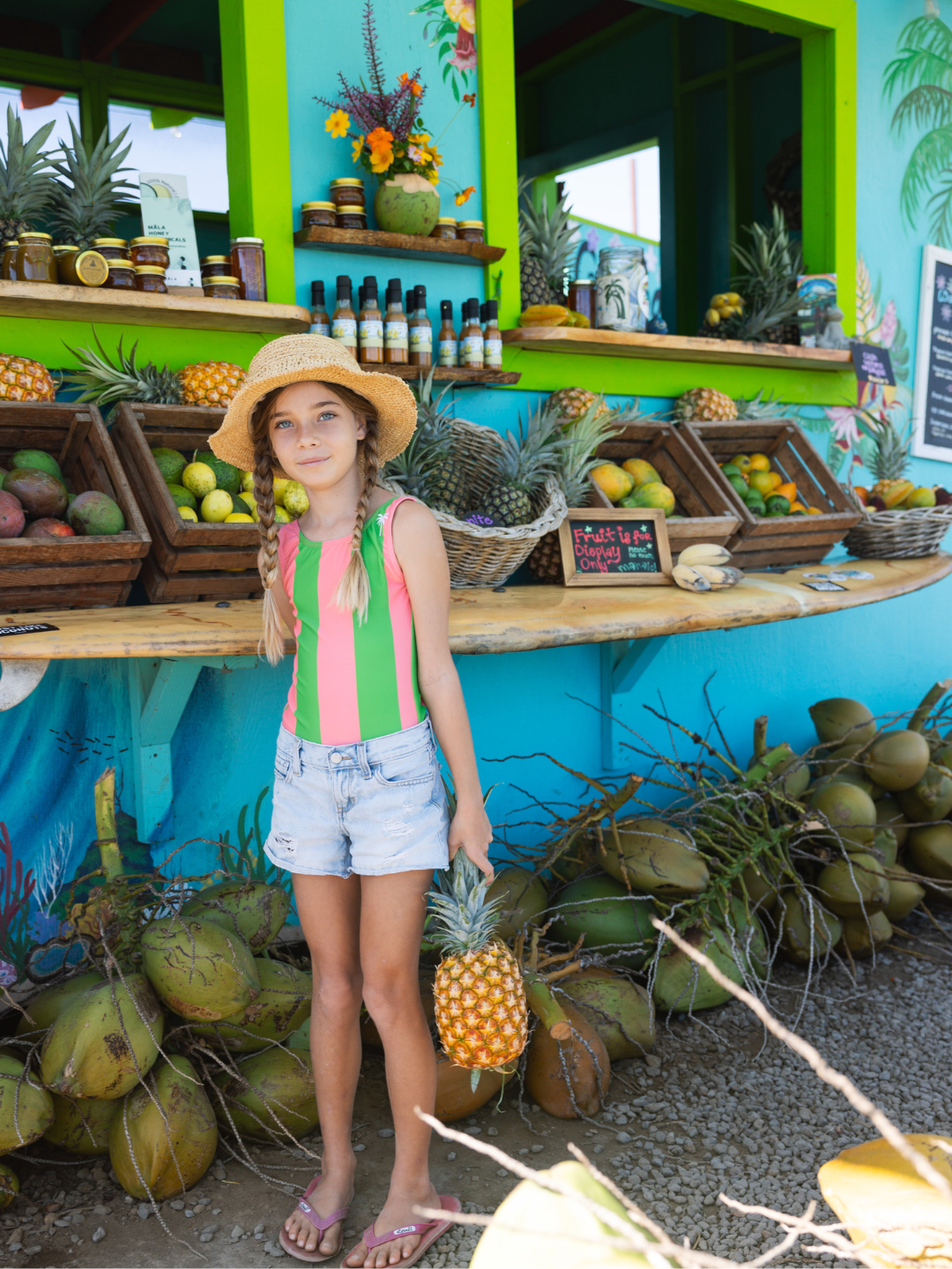 Watermelon | A child is wearing Retro Stripe Swimsuit in Watermelon with shorts at the fruit market holding a pineapple. 