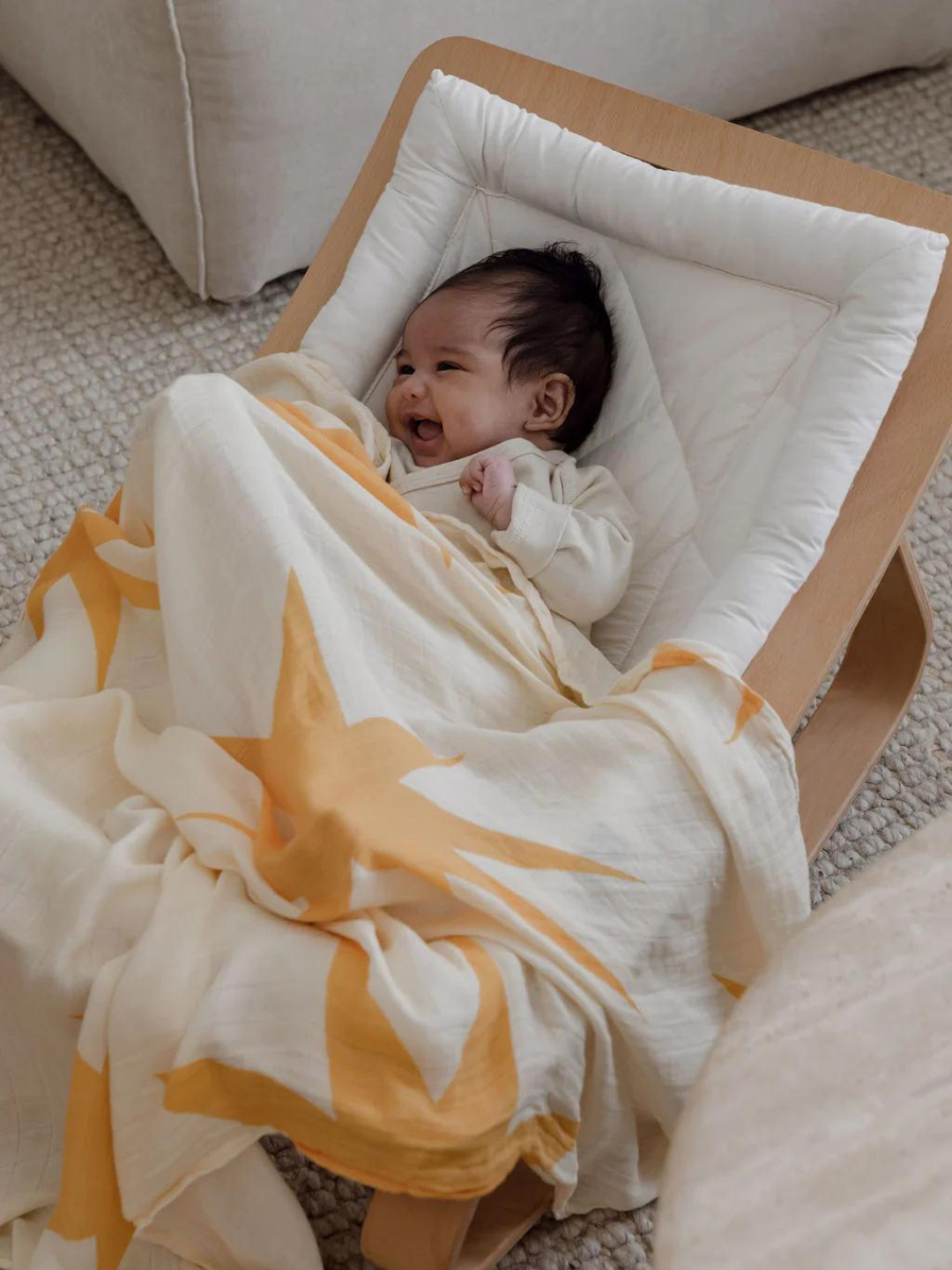 A baby in a white cushioned chair, smiling and looking upward, is wrapped in soft ORGANIC BAMBOO SWADDLE WRAP and covered with a yellow star-patterned blanket.