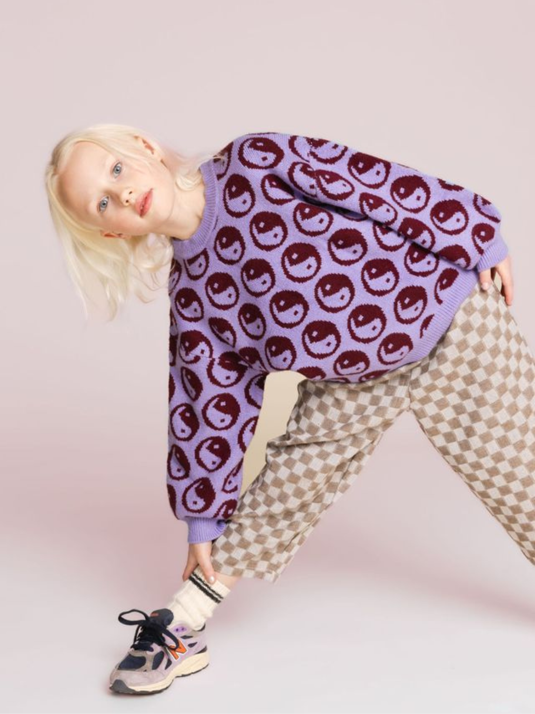 Violet | A child with blonde hair is wearing the COSMOS SWEATER, a super-soft purple crewneck adorned with smiley faces, paired with checkered pants. The child is bending forward slightly while looking at the camera against a plain background.