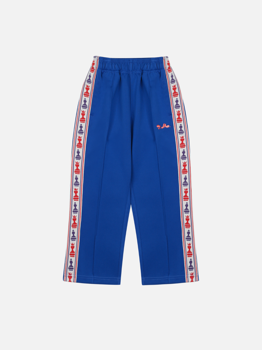 Image of Blue | The CHESS TRACK PANTS in blue feature an elastic waistband, red, white, and blue logo tape along the side seams, and a small red logo on the left thigh; laid flat on a white background for a comfortable and stylish look.