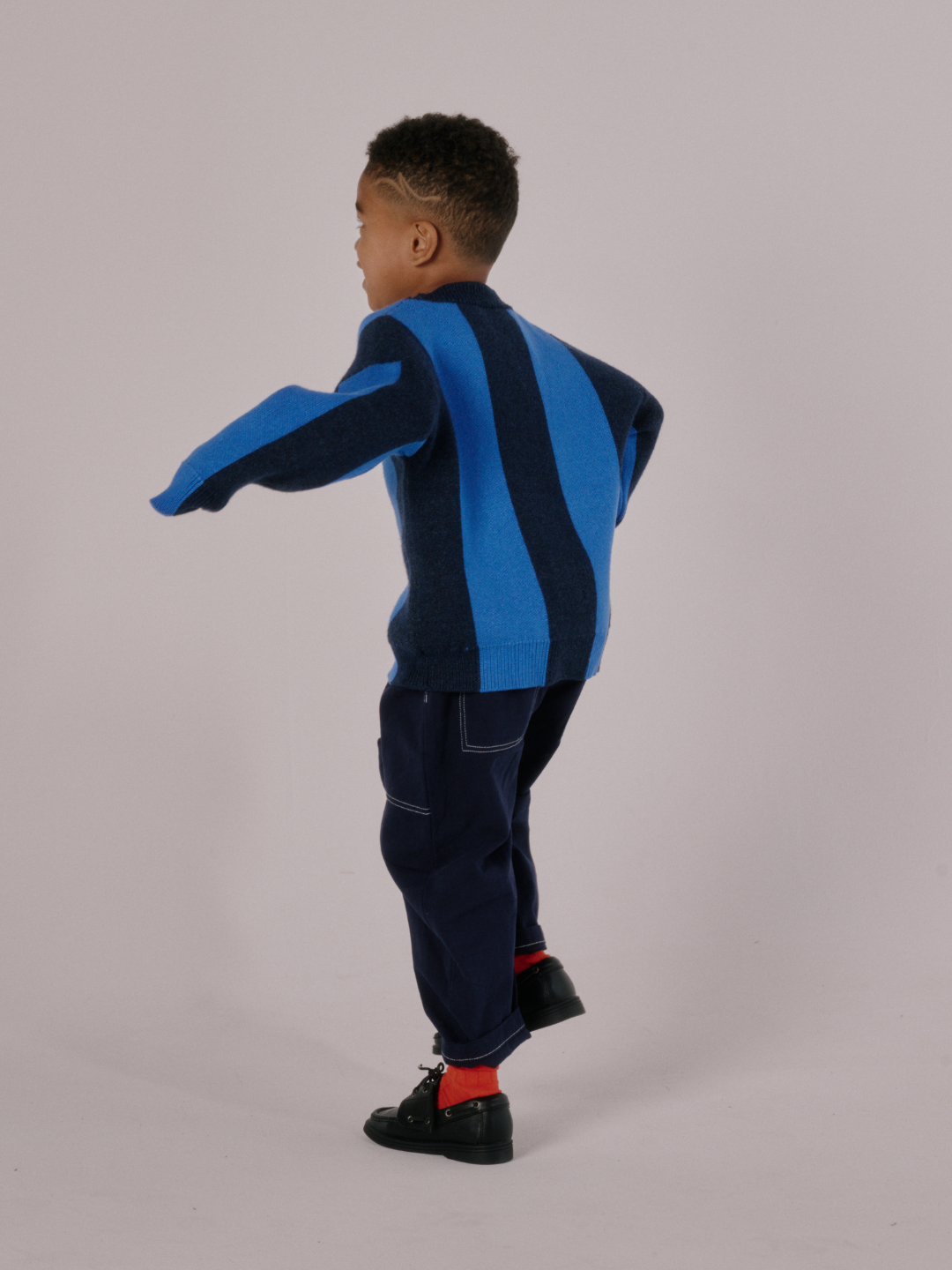 A young child stands sideways against a plain background, wearing the STRIPE CARDIGAN paired with navy pants, red socks, and black shoes, arms slightly raised.