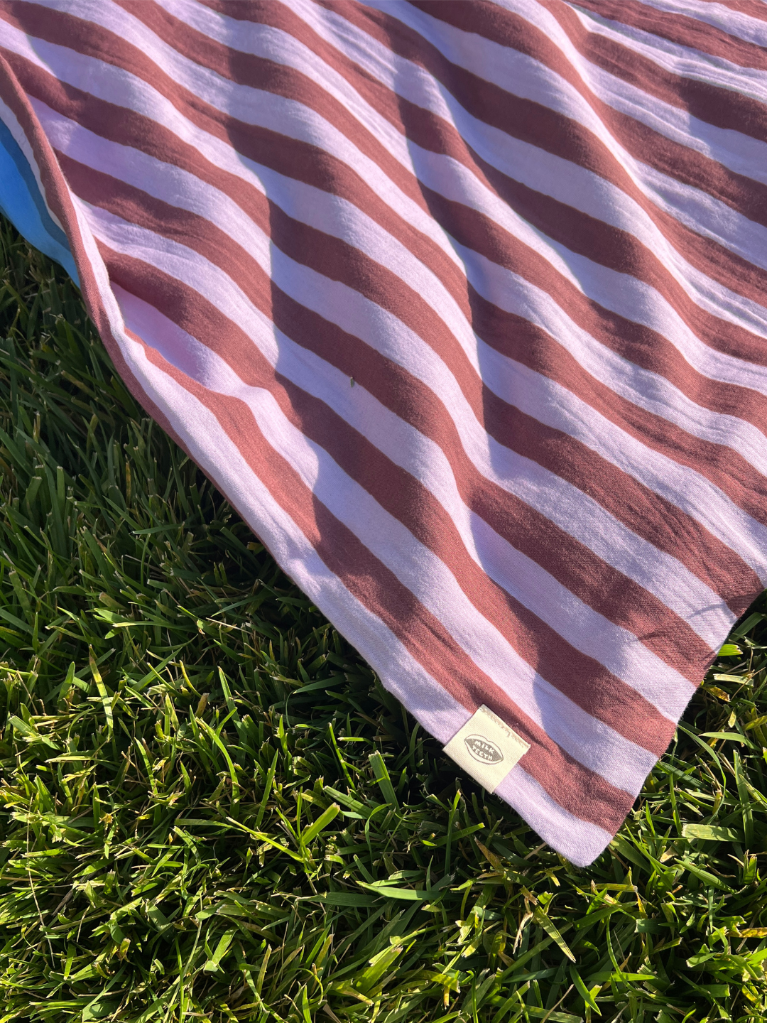 Detail image showing the corner or the organic cotton swaddle in purple and maroon stripes, on grass, with a cream Milk Teeth logo tag.