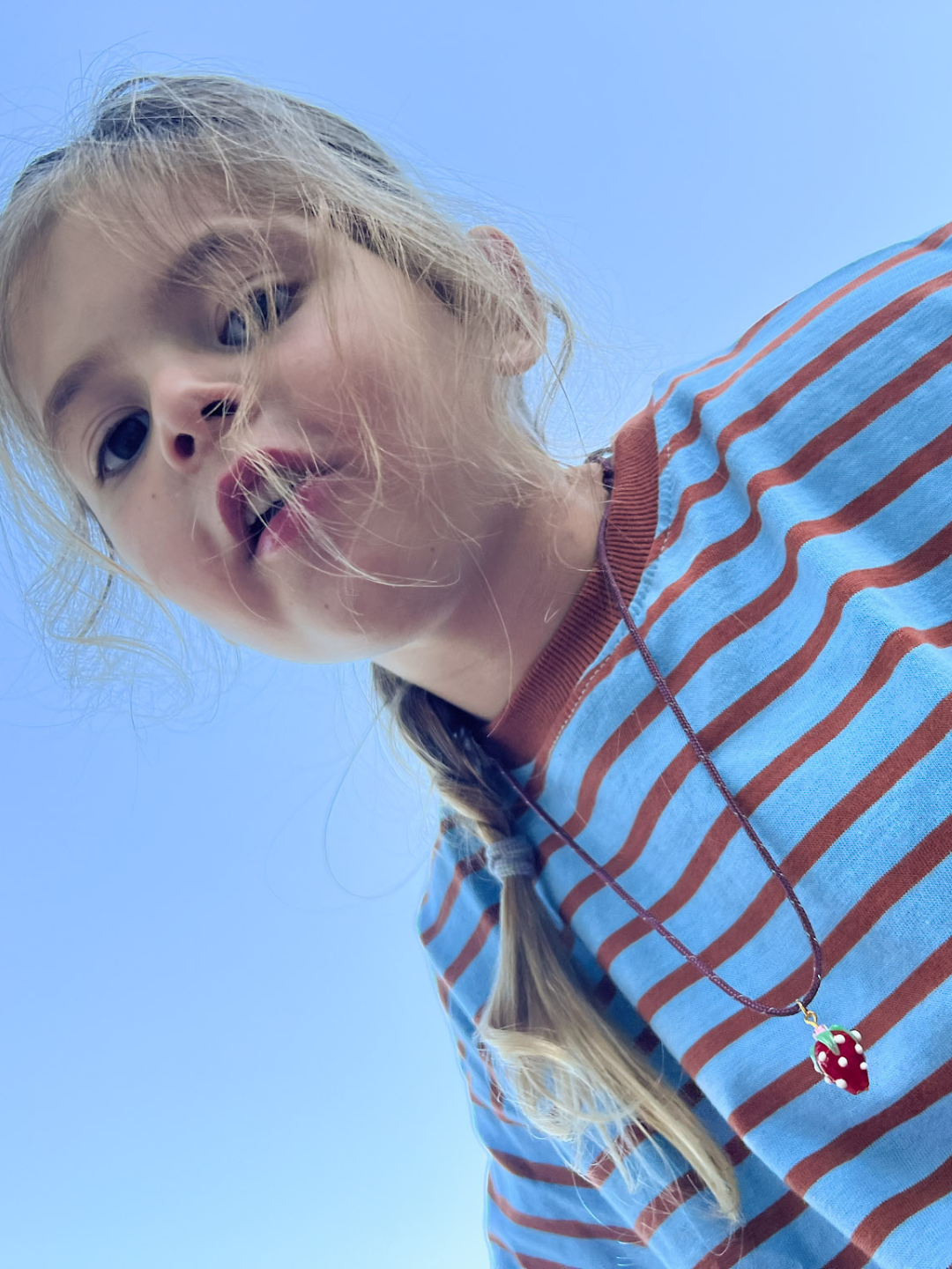 A young girl with braided hair wears a blue and brown striped shirt and the FRAGOLA NECKLACE, a handmade glass strawberry charm, as she looks down at the camera against a clear blue sky.