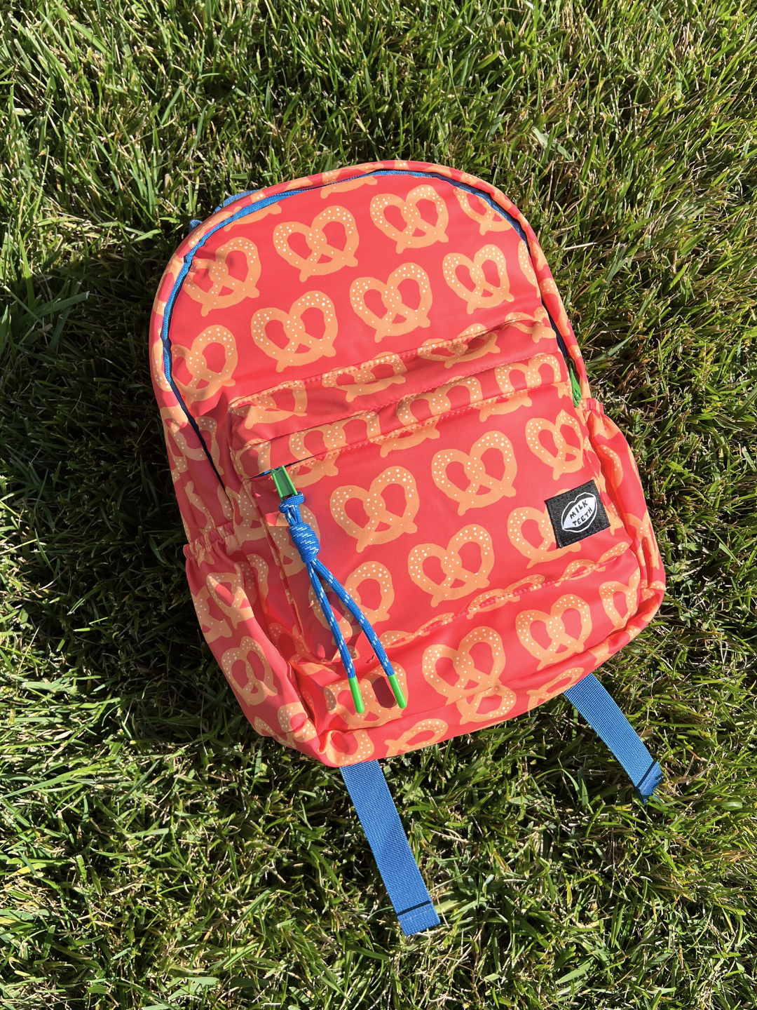 A red and tan backpack with a repeating pretzel pattern, featuring blue accents on the zipper pulls, top handle, and piping, and a black Milk Teeth logo patch on the front pocket, shown lying on green grass.