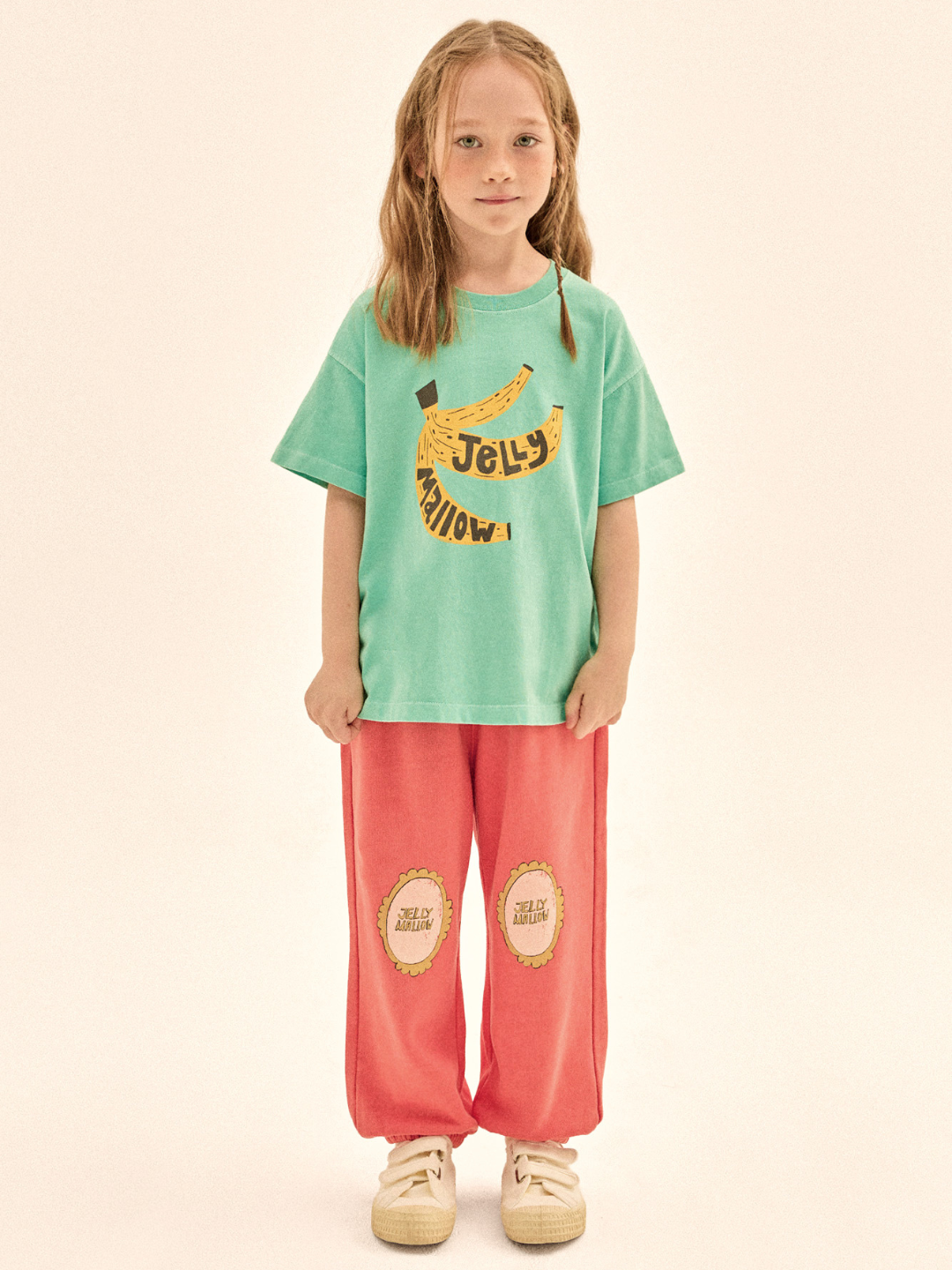 Child wearing a green t-shirt with a banana graphic and red pants on a beige background