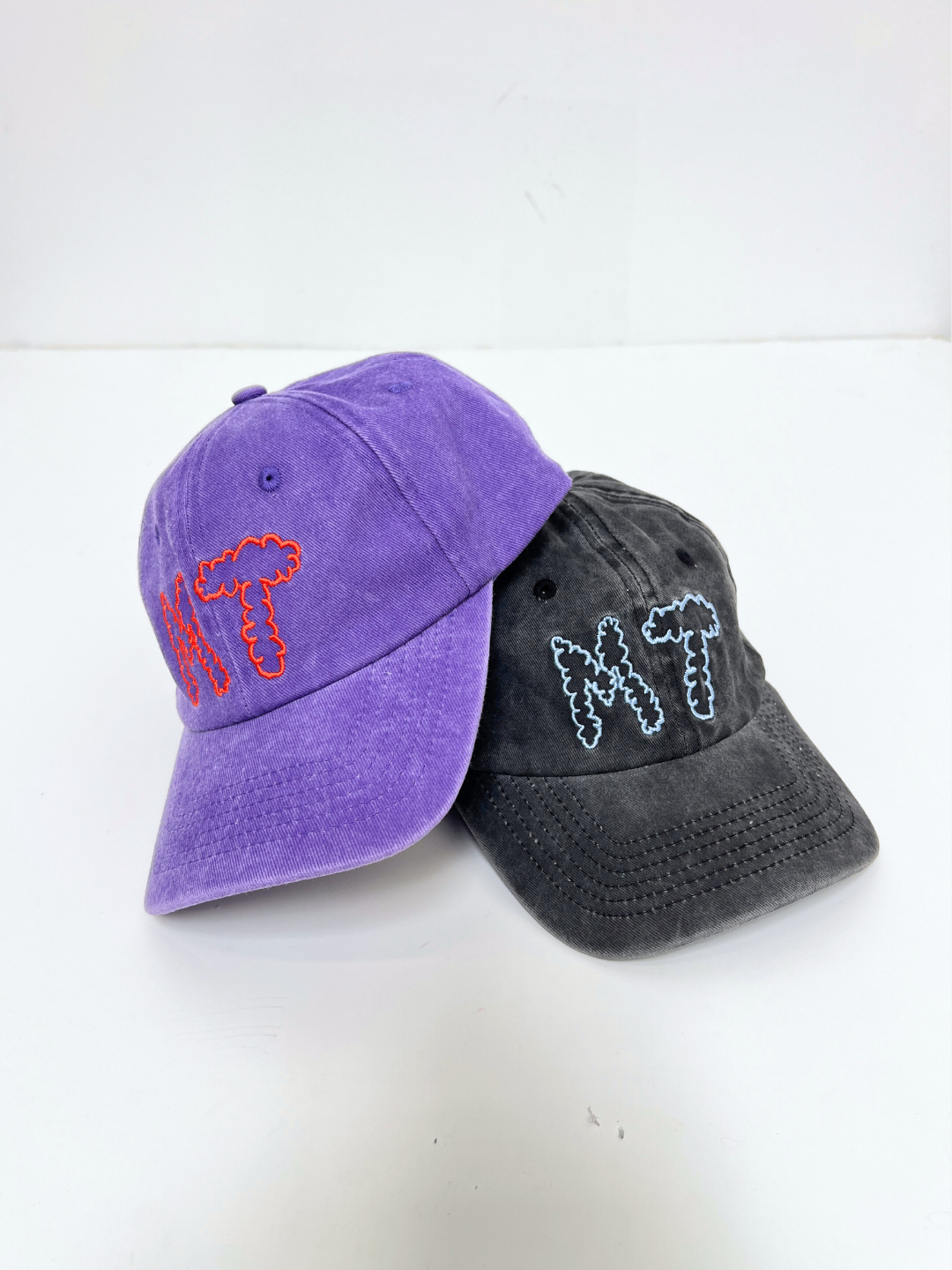 Two MT DAD CAPs, one purple with red embroidered "MT" and one black with white and blue "MT," made from cotton canvas, are shown on a white surface.