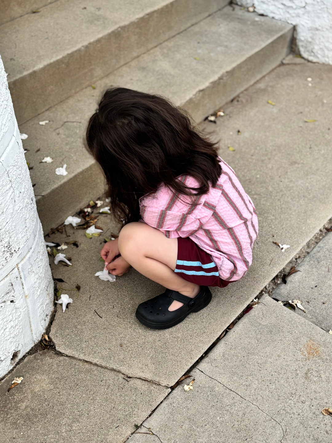 Maroon | A child with long dark hair, wearing a pink striped shirt and SPRINTER SHORTS with an elasticated waist, squats near steps, picking up white flower petals from the ground.