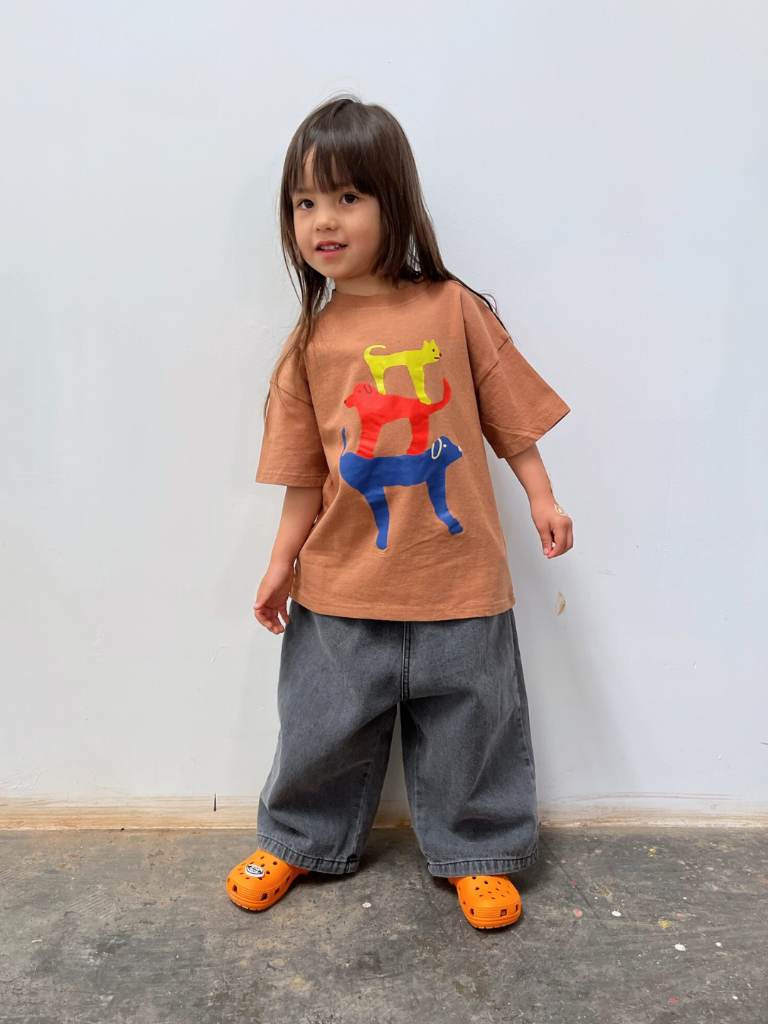 A young child with long hair wears a brown t-shirt with playful animal prints, PEPPER JEAN wide gray jeans, and bright orange crocs, standing against a simple white wall.