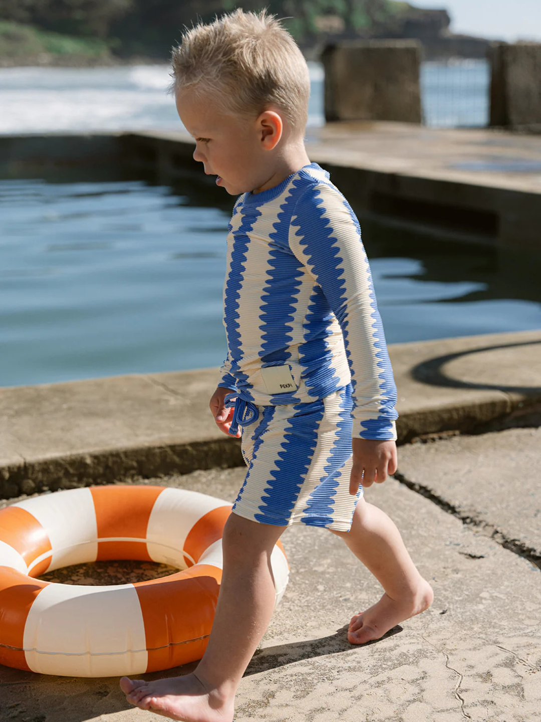 A young child wearing blue and white zigzag-patterned SWIM SHORTS with UV protection walks barefoot near a lifebuoy by a pool.
