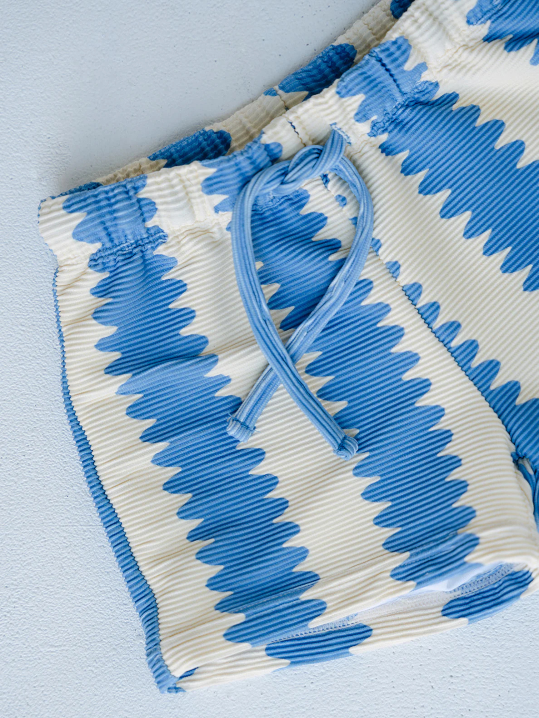Close-up of SWIM SHORTS featuring a blue and white zigzag pattern, elastic waistband, and drawstring, made from recycled material and displayed on a light-colored surface.