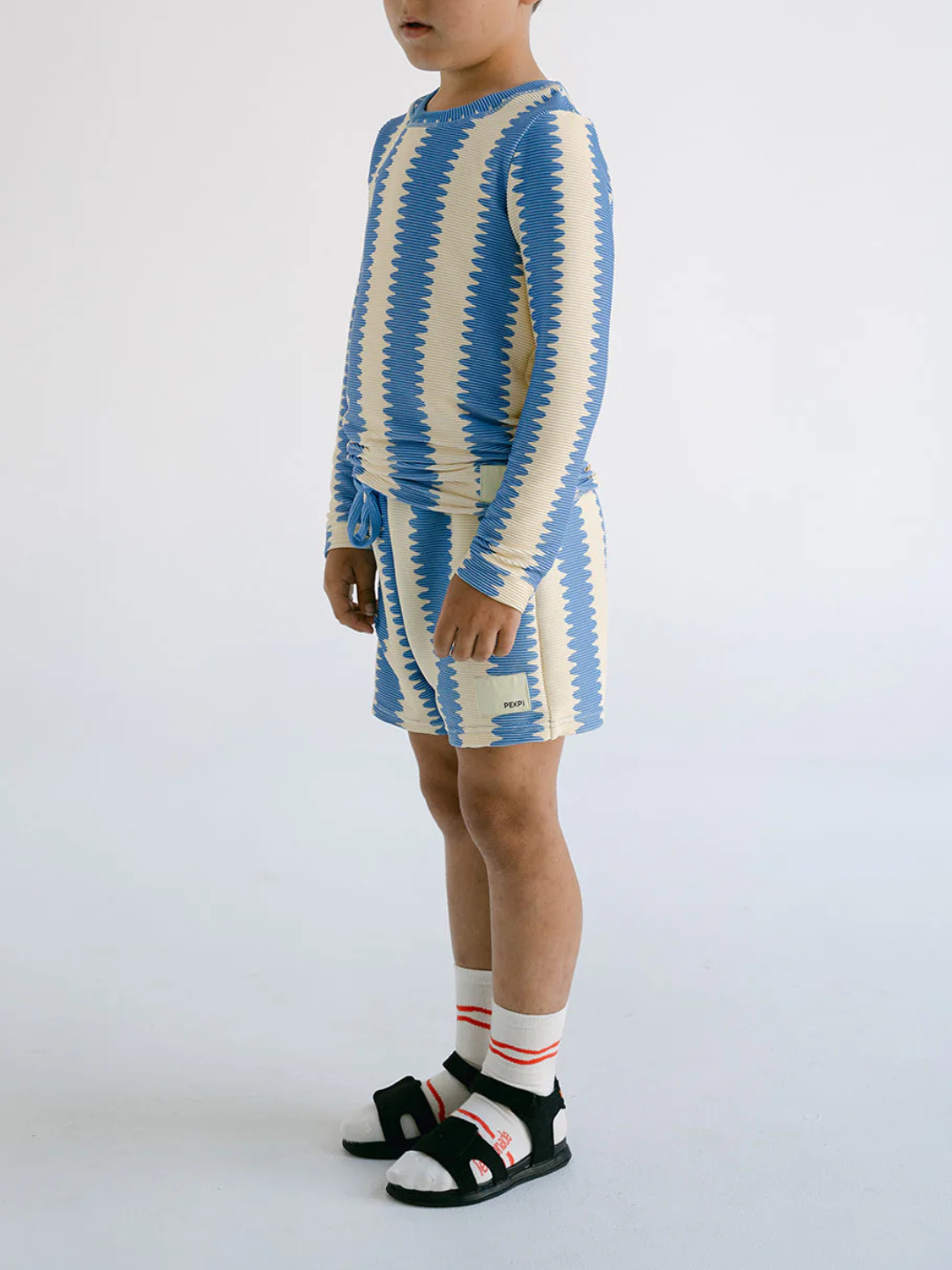 A child wears SWIM SHORTS in a blue and beige zigzag pattern made from recycled material, paired with a matching long-sleeve shirt, white socks with red stripes, and black sandals against a plain background.