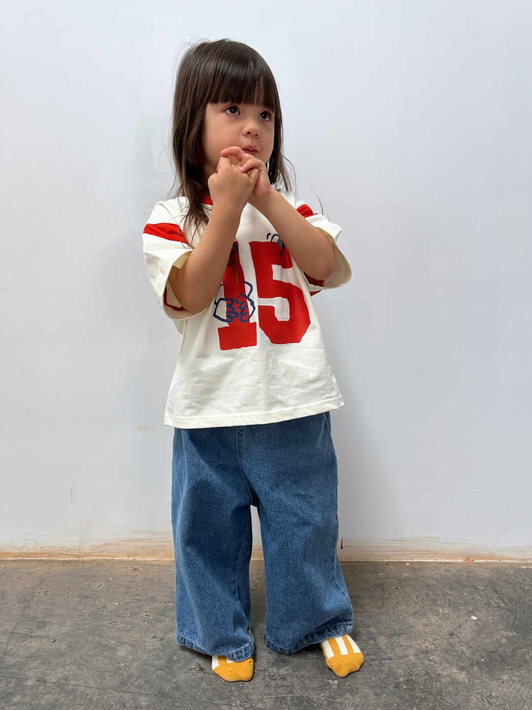 Blue | A young child with long brown hair stands indoors, wearing the PEPPER JEAN blue jeans, a white shirt with red numbers, and yellow shoes, hands clasped near the face.