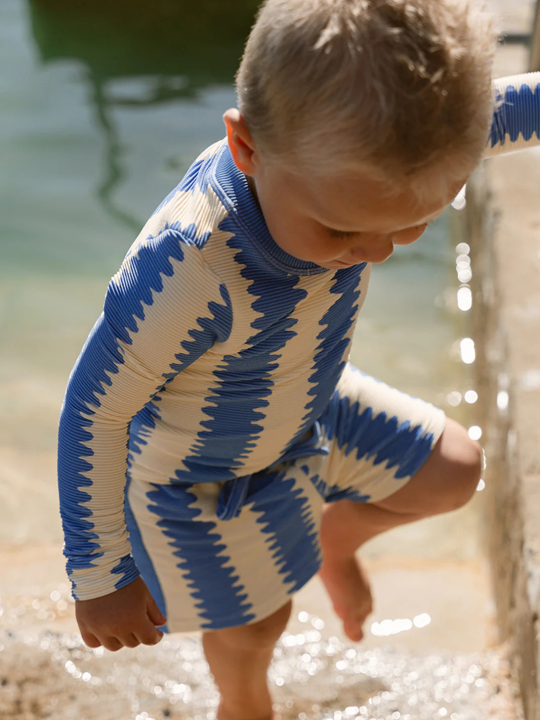 A young child in SWIM SHORTS with blue and beige zigzag patterns and UV protection steps from shallow water onto a stone edge, sunlight catching their hair and outfit.