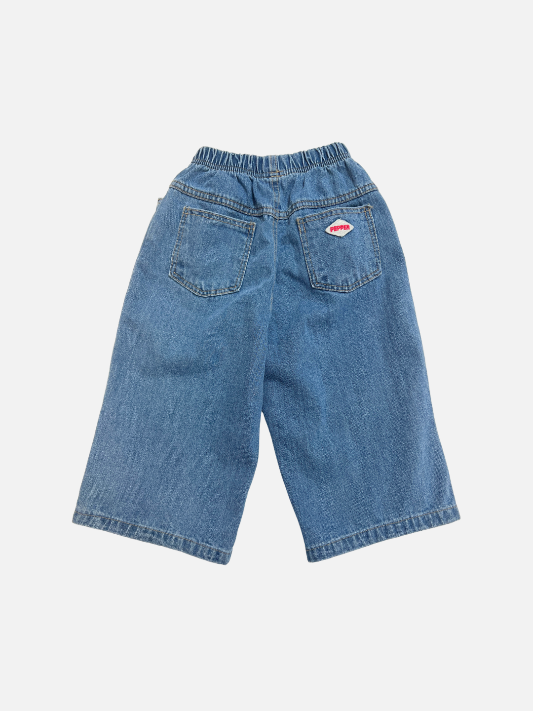 The PEPPER JEAN features loose-fitting blue denim shorts with an elastic waistband and two back pockets, one adorned with a small red and white patch.