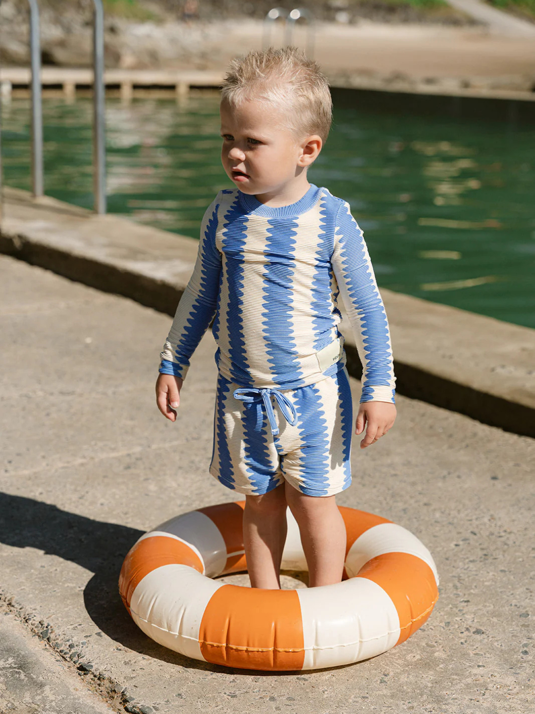 A young child wearing SWIM SHORTS stands inside an orange and white inflatable swim ring on a concrete surface near water.