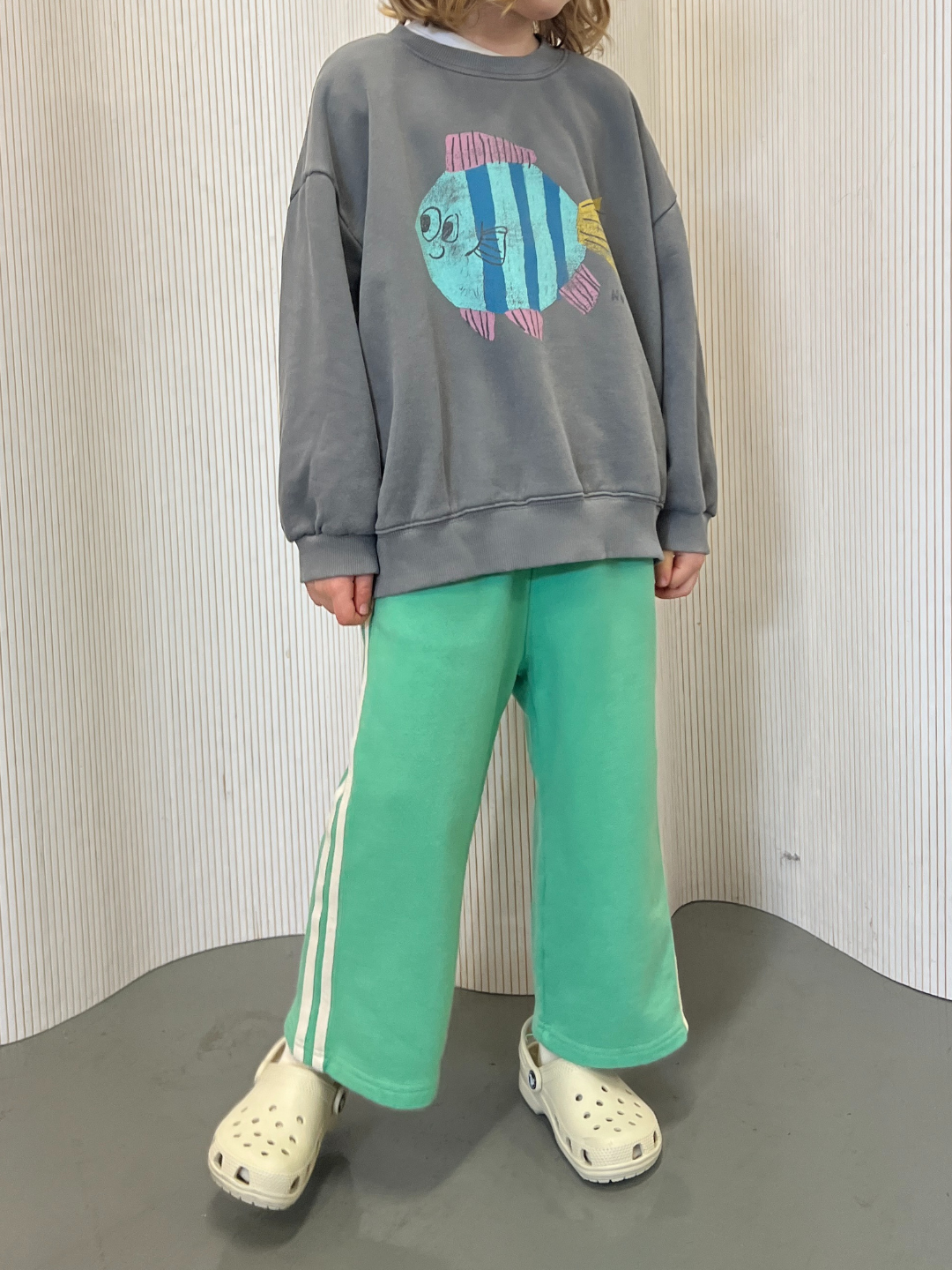 A person stands against a striped backdrop, wearing a gray fish-printed sweatshirt made from cotton loop-back terry, paired with MINTY STRIPE TRACK PANTS in mint green with white stripes, and completed with beige clogs.