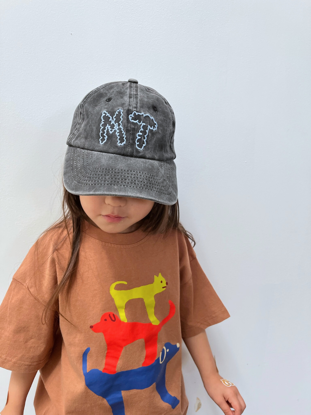 A child wears a brown T-shirt with colorful stacked animals and the MT DAD CAP, a gray cotton canvas baseball cap featuring "MT" for Milk Teeth, standing against a plain light background.