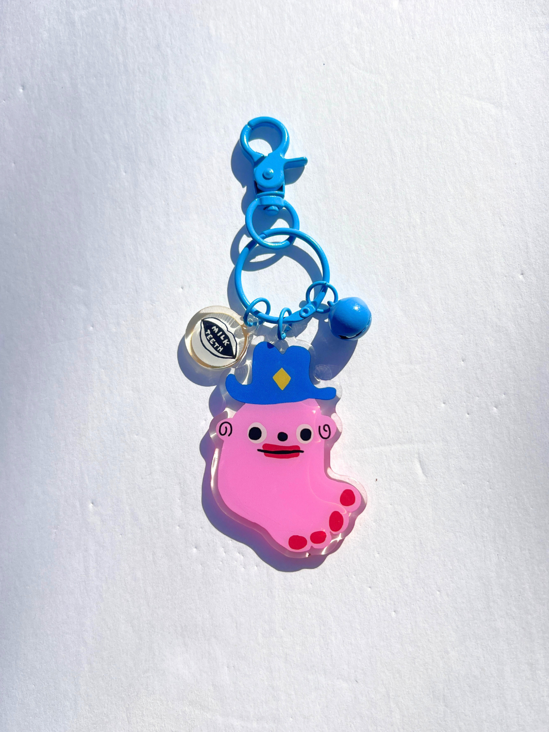 Foot | The SOUVENIR KEYCHAIN by Katie Kimmel features a pink cartoon foot with a blue hat, blue clasp, and a round tag—ideal as a quirky keychain charm or fun bag accessory, displayed on a white background.