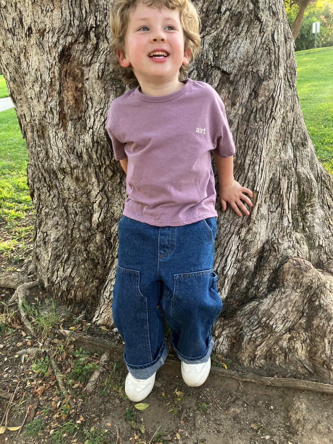 A child is wearing the Studio Tee in Mauve with dark denim jeans and white sneakers, standing next to a tree