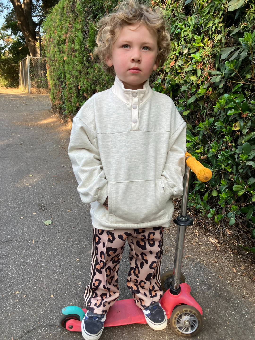 A child stands confidently on a red scooter along the sidewalk, dressed in a cozy EVERYDAY PULLOVER in heather grey and stylish leopard print pants.