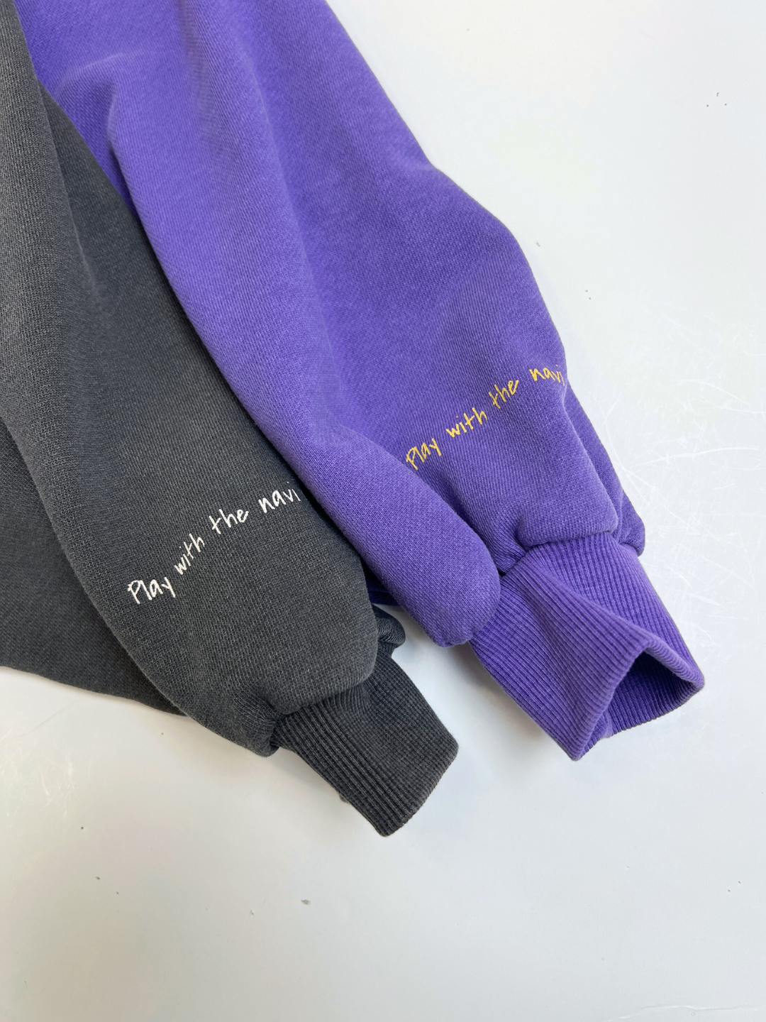 Purple | Two sleeves—one black, one purple—from the PLAYTIME SWEATSHIRT show “Play with the wave.” embroidered near the cuff in white (on black) and yellow (on purple) thread, part of a Korean kids sweatshirt collection.