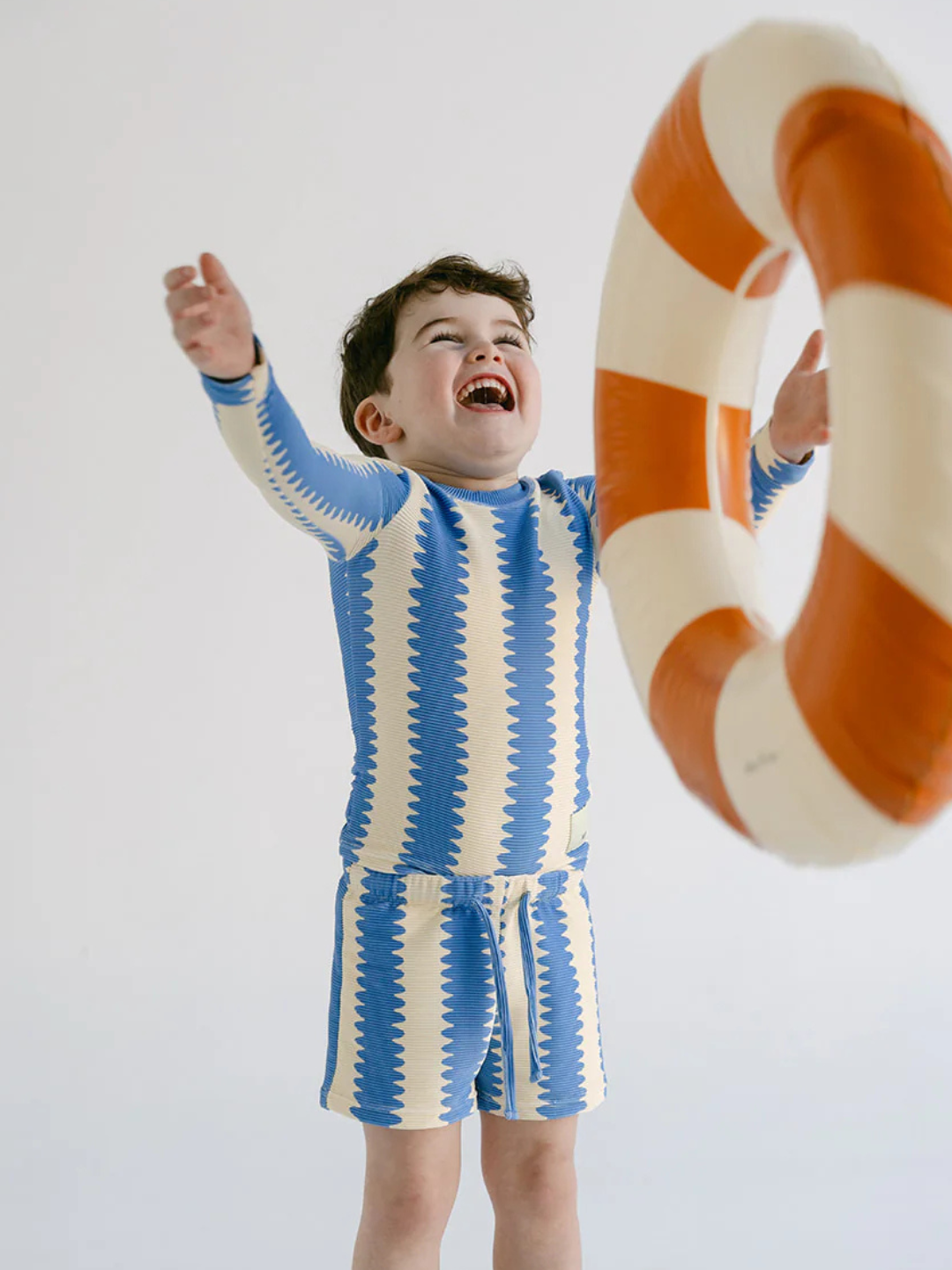 A young child smiles with arms raised, reaching toward an orange and white inflatable swim ring while wearing blue and white patterned SWIM SHORTS.