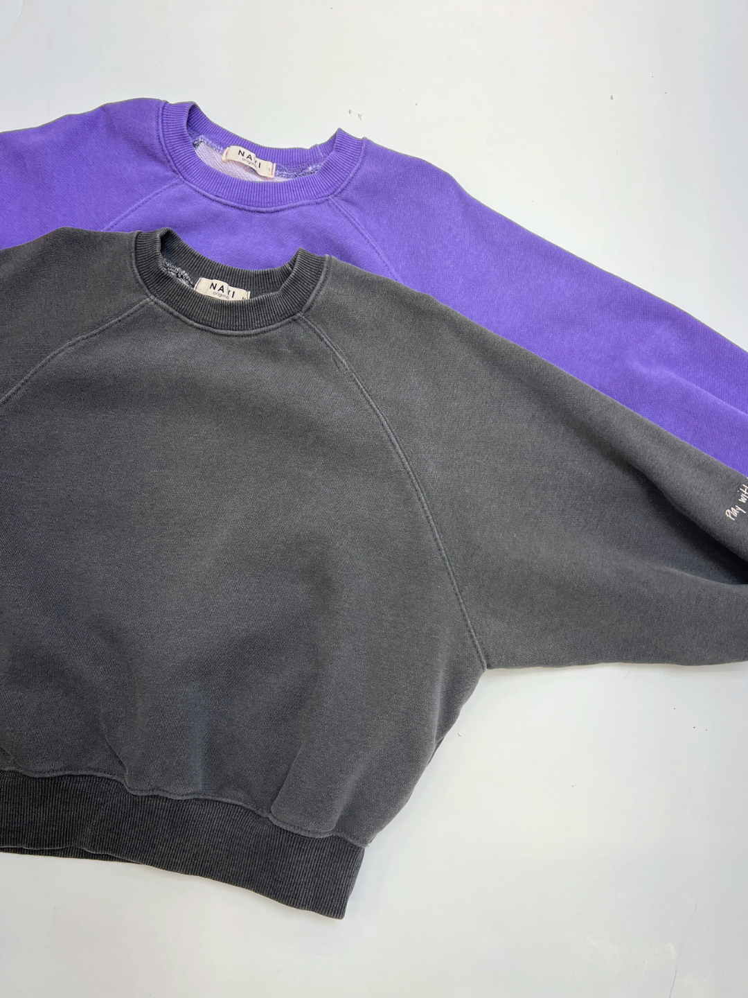 Two PLAYTIME SWEATSHIRTS for kids, one purple and one dark gray, are shown laid flat with overlapping sleeves. Made from soft garment-dyed cotton, these Korean crewnecks blend style and comfort.