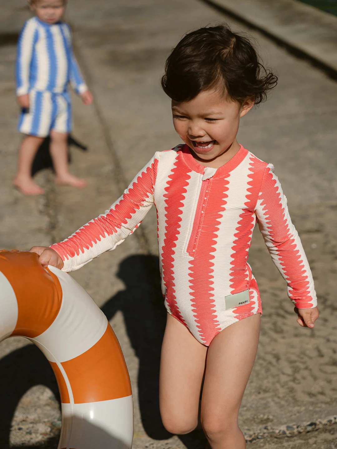 A young child in a red and white FULL PIECE SWIMSUIT with UPF 50+ UV protection holds a life preserver, while another child in a blue and white FULL PIECE SWIMSUIT stands nearby by the water.