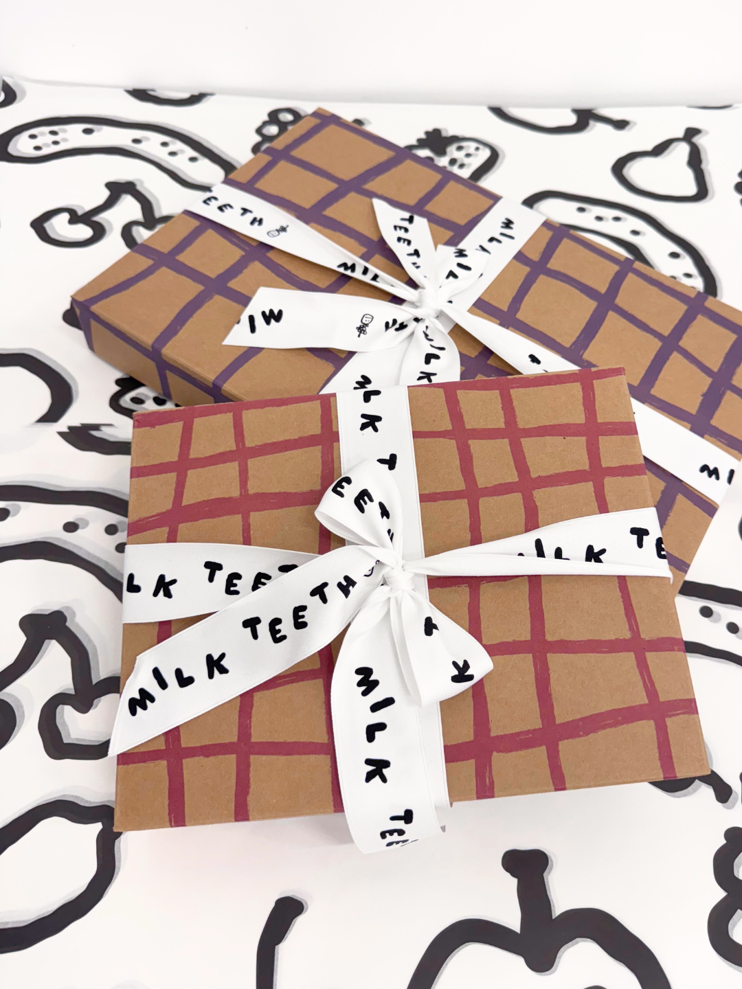 Gift Boxes with plaid brushstrokes wrapping and 'Milk Teeth' ribbon on a patterned background