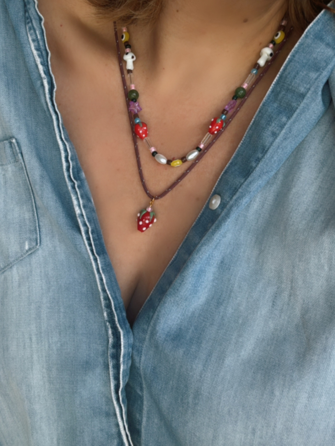 A person wearing a blue denim shirt and two beaded necklaces, including the FRAGOLA NECKLACE from Meyous jewelry with colorful beads, mushroom pendants, and a handmade glass strawberry charm.