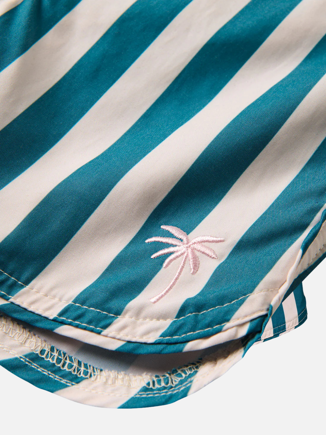 Close-up of the RETRO FLARE BOARDSHORTS CABANA STRIPE: teal and white striped recycled polyester fabric with a small pink embroidered palm tree near the scalloped hem—perfect for stylish swimwear.