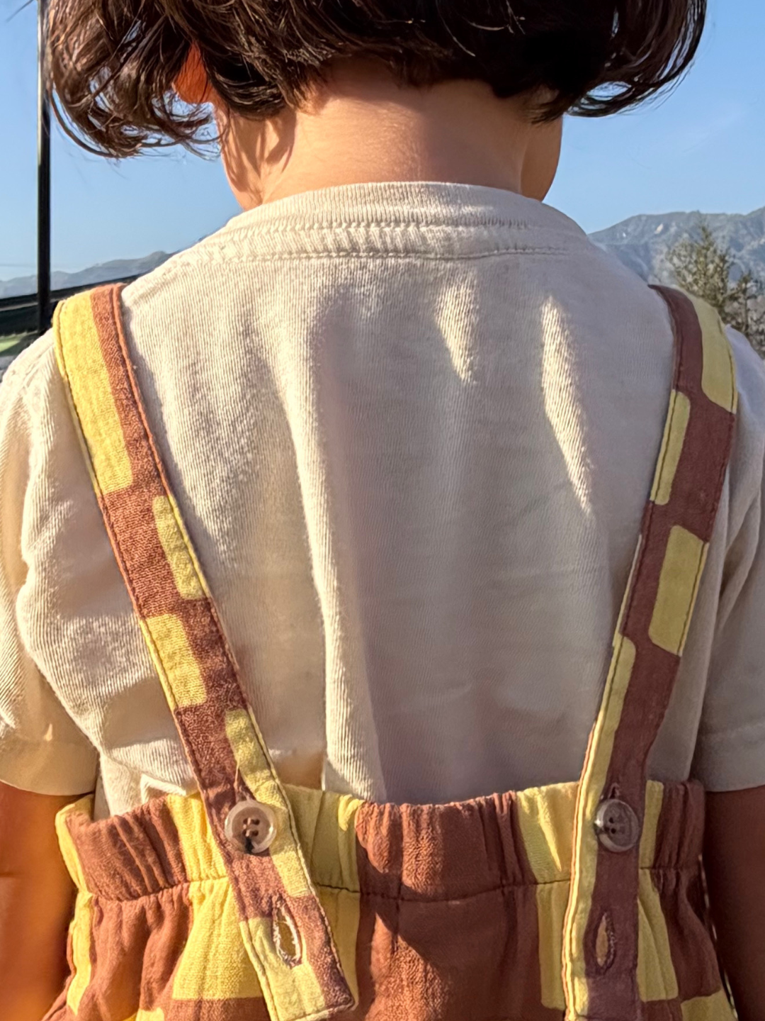 Yellow Checkers | Child wearing a white shirt and brown overalls with yellow accents, standing outdoors.