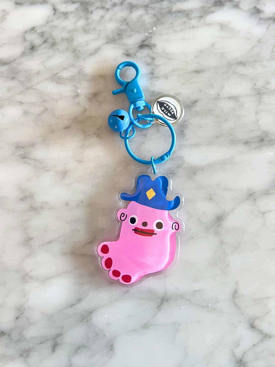 Foot | The SOUVENIR KEYCHAIN features a pink cartoon foot character in a blue hat, designed by Katie Kimmel, shown on a marble surface—an ideal playful accessory for your bag.