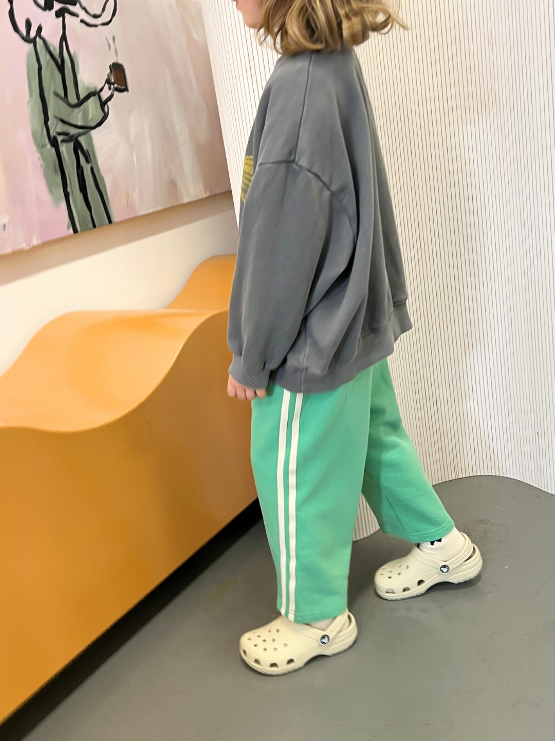 A child in a gray sweatshirt and Bon Bon Butik's MINTY STRIPE TRACK PANTS stands on white clogs in a room with abstract art and an orange wave-shaped bench.