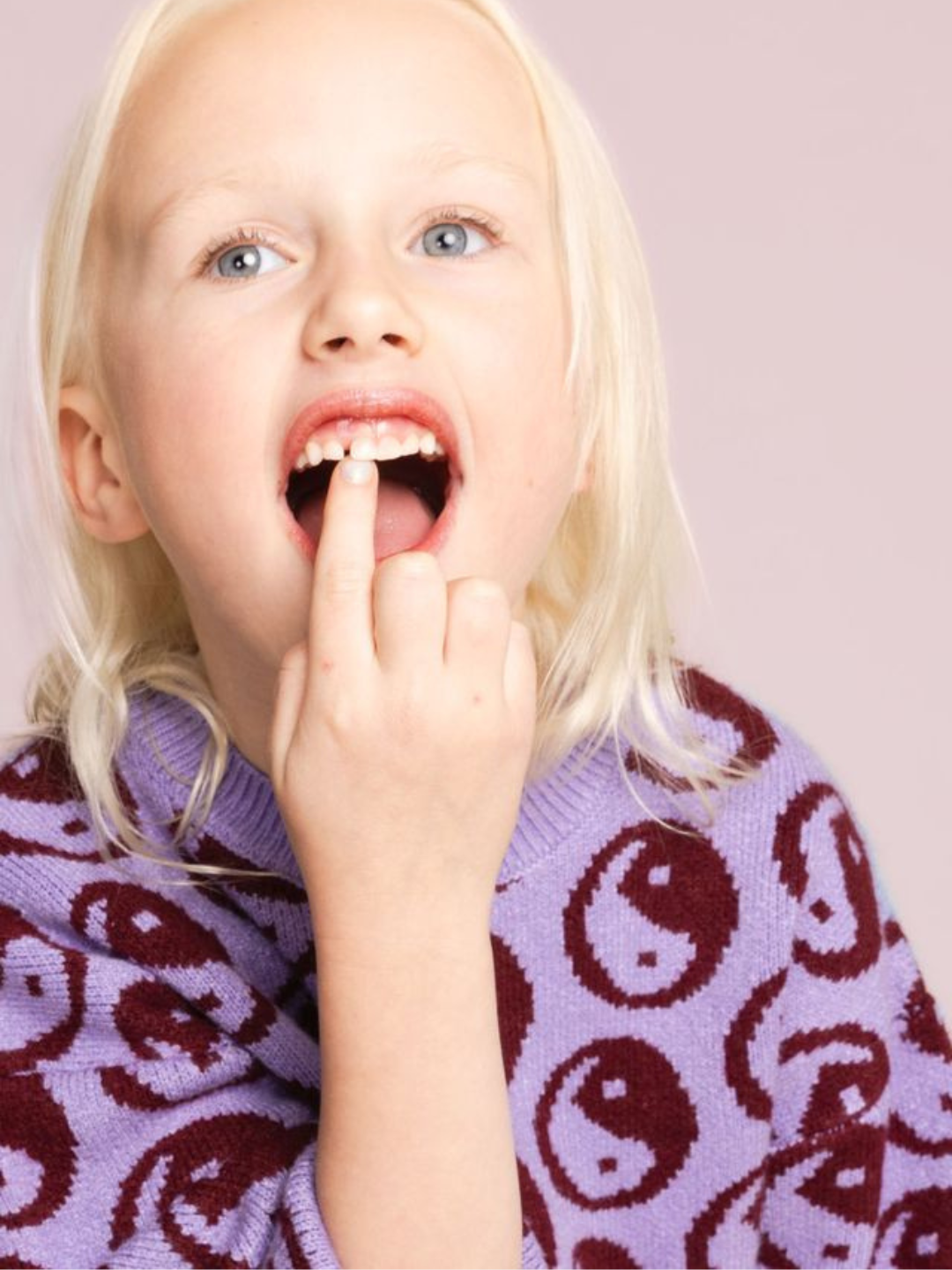 A child with blonde hair wearing a COSMOS SWEATER, a super-soft purple crewneck with a yin-yang intarsia knit pattern, points to a missing tooth in their mouth.