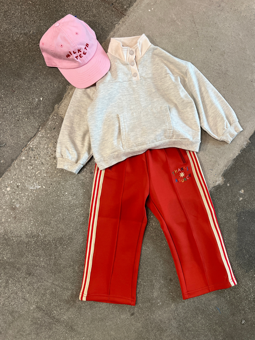 An EVERYDAY PULLOVER in heather grey, orange pants with white stripes, and a pink cap featuring text are set against a concrete background.