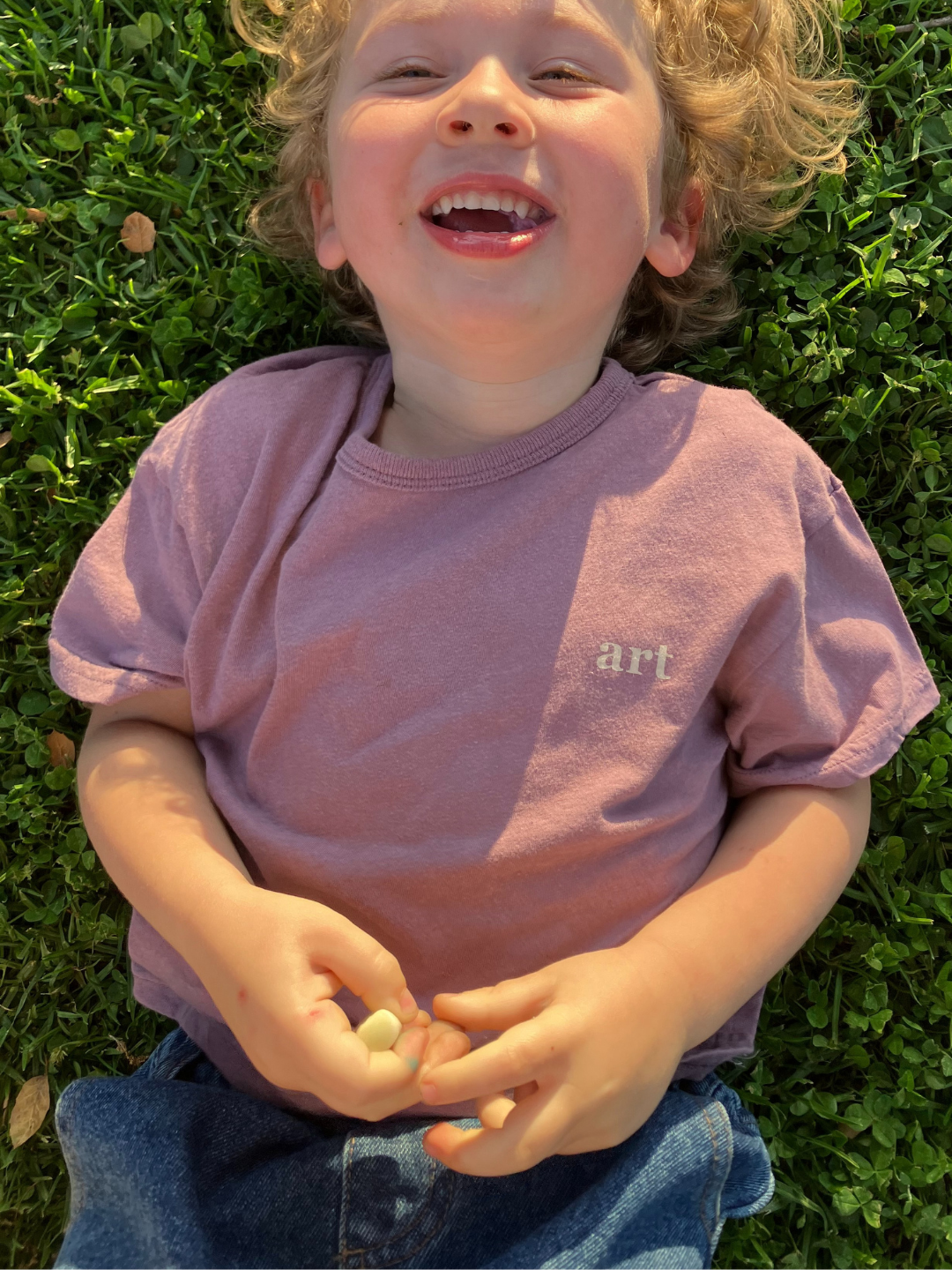 Mauve | A child is wearing the Studio Tee in Mauve with dark denim jeans, laying on the grass laughing