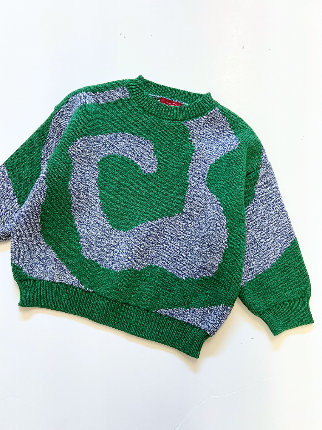 The SWIRL SWEATER, featuring green and gray abstract swirl patterns, is laid flat on a white background.