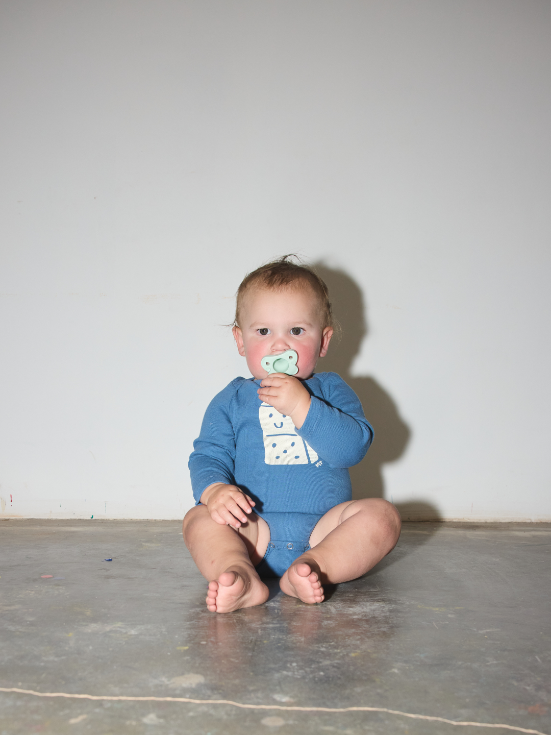 A baby sitting on a concrete floor against a plain wall, wearing a blue long-sleeve bodysuit with a white graphic print on the front and holding a pacifier.