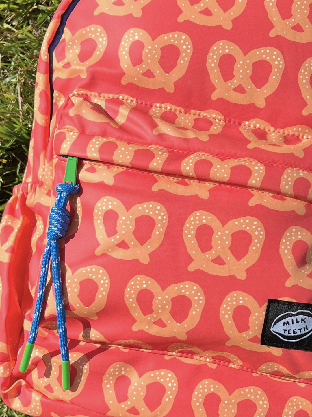 A close-up of a red backpack with a repeating pretzel pattern, featuring a blue and green zipper pull and a black 'Milk Teeth' brand patch.