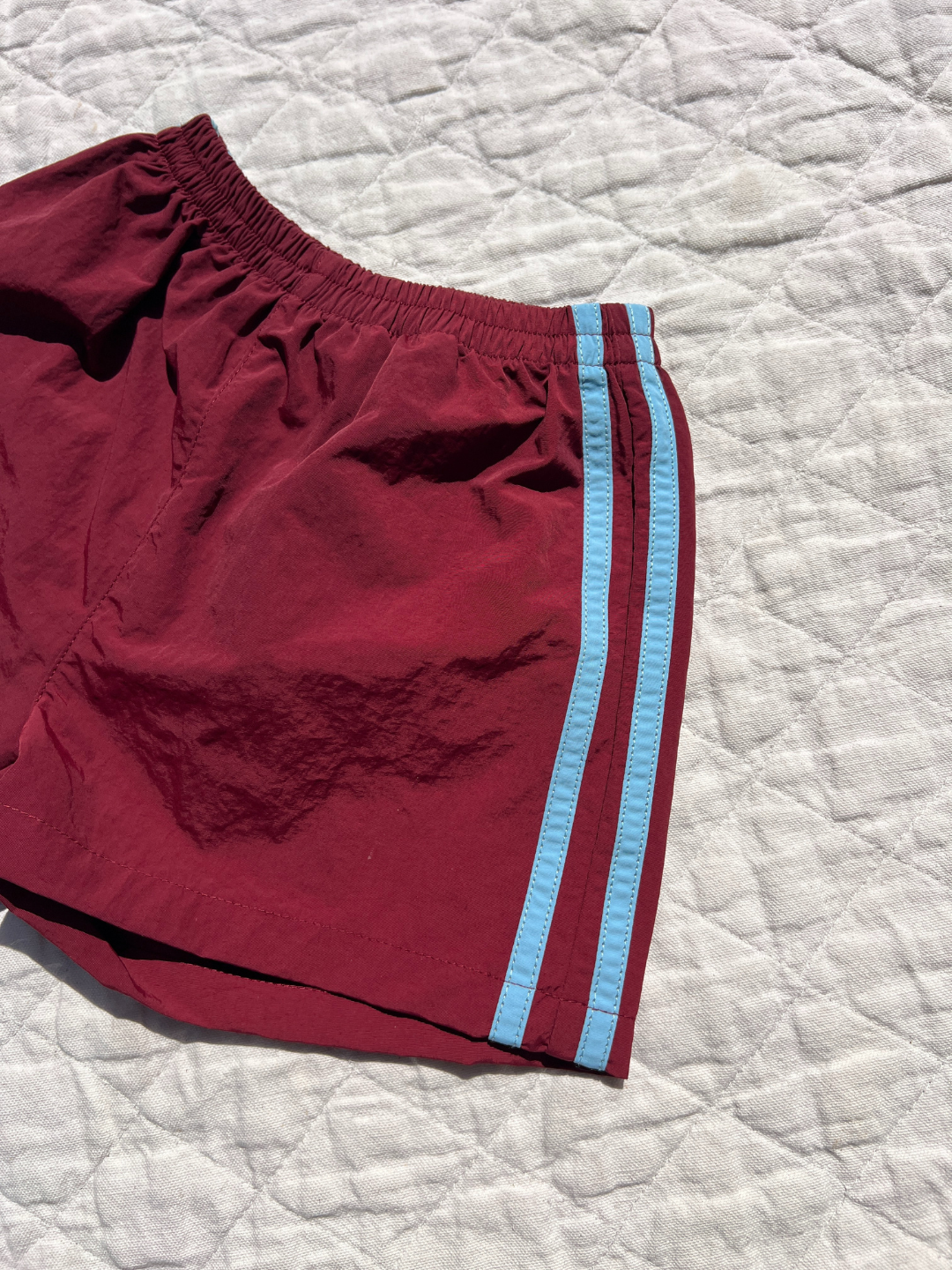 A pair of maroon SPRINTER SHORTS with an elasticated waist and two light blue side stripes is laid flat on a textured white quilt.