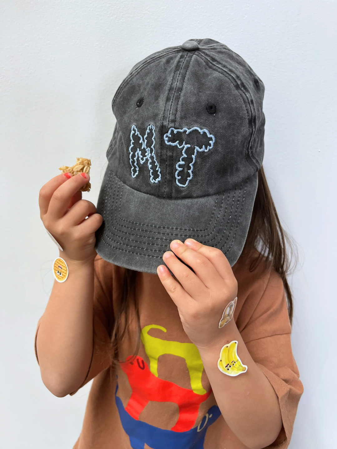 Charcoal | A child wearing the MT DAD CAP, a gray cotton canvas baseball cap with "MT" embroidered, holds a snack while covering part of their face; stickers decorate their hands, arm, and shirt.