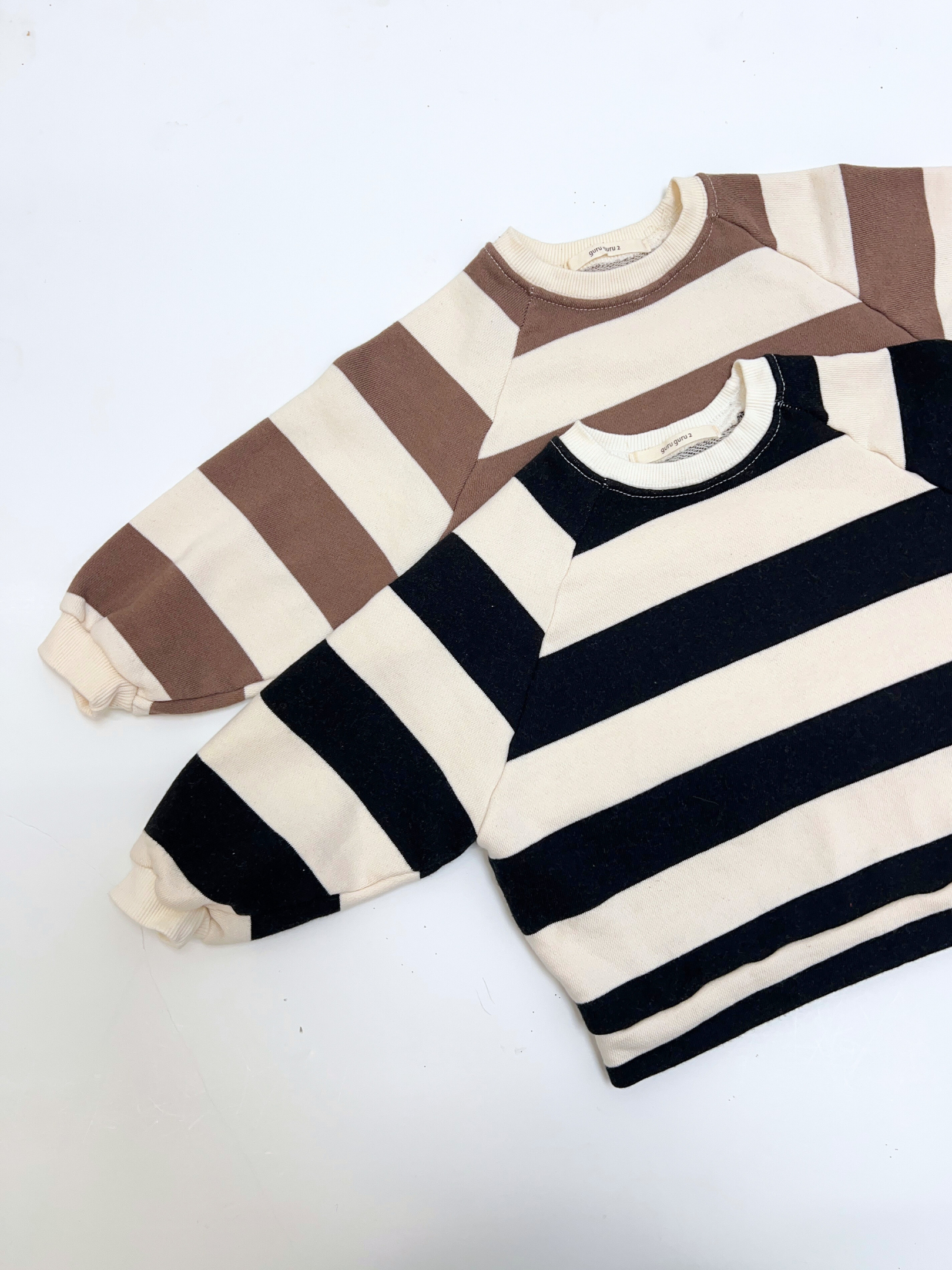 Two STRIPE BABY SWEATSHIRT tops from a Korean brand, made of soft cotton terry in wide stripes—one brown and cream, one black and cream—are shown with long sleeves, laid flat on a white background.