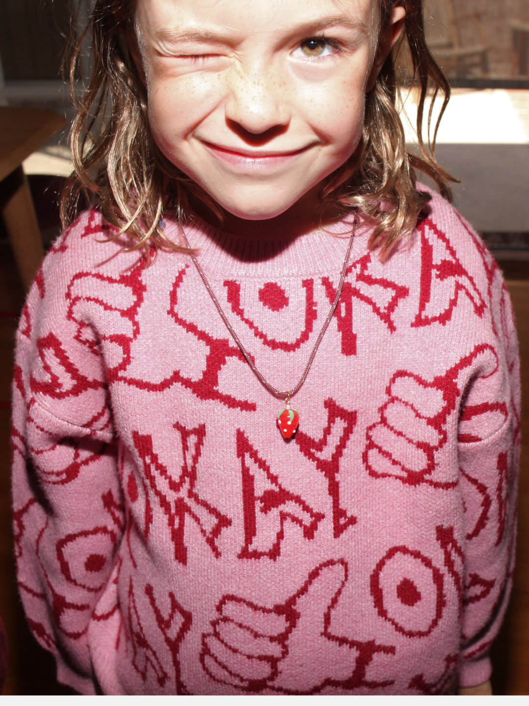A child with wet hair winks and smiles in a pink sweater with "OKAY" text, wearing the FRAGOLA NECKLACE by Meyous jewelry, featuring a handmade glass strawberry charm.