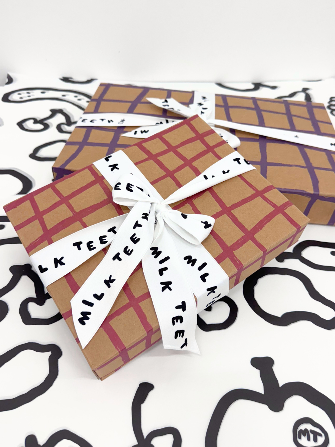Gift Boxes with plaid brushstrokes wrapping and 'Milk Teeth' ribbon on a patterned background