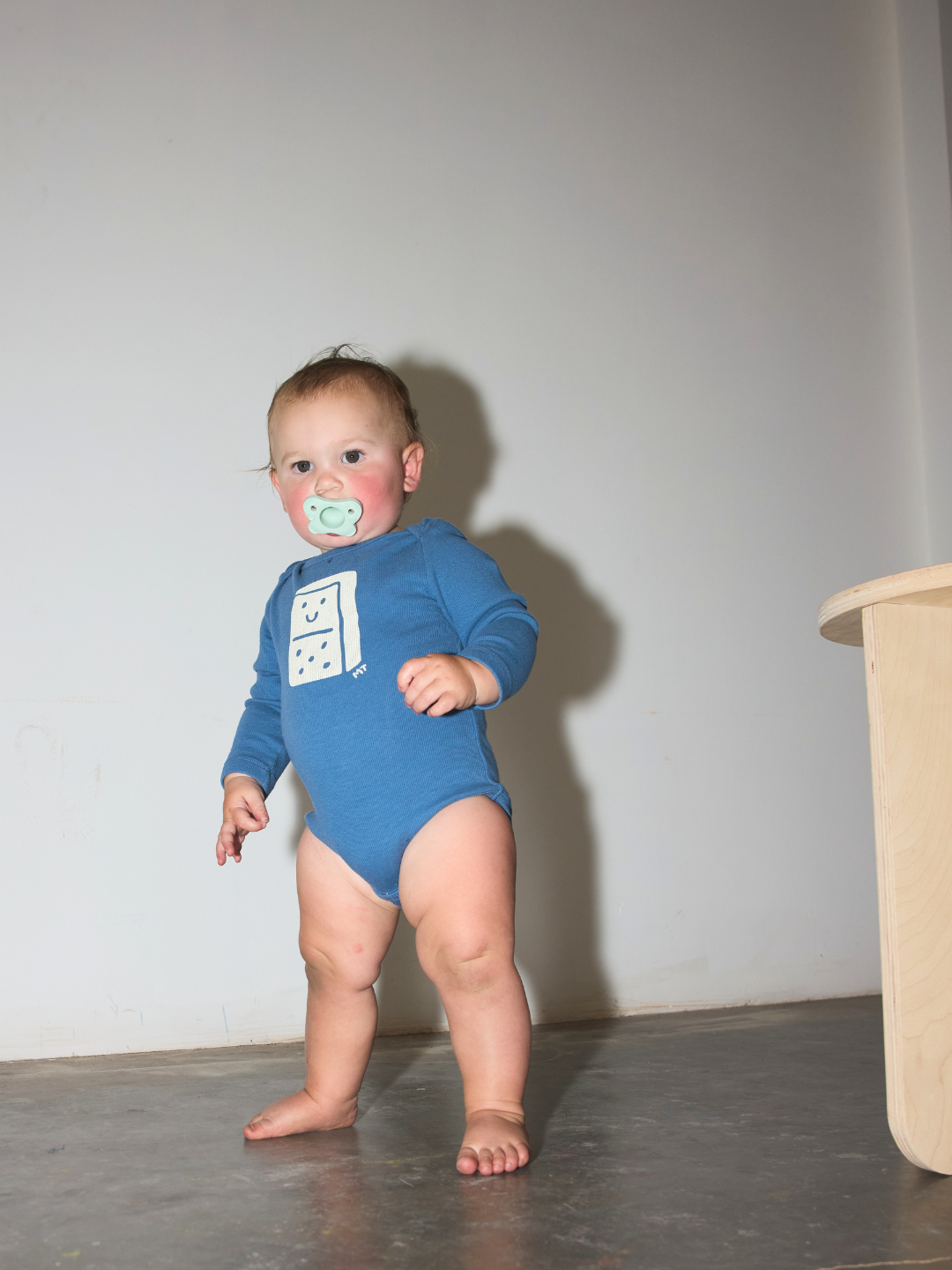 A baby standing barefoot indoors, wearing a blue long-sleeve bodysuit with a white graphic print on the chest and a pacifier in their mouth, against a plain light gray wall.