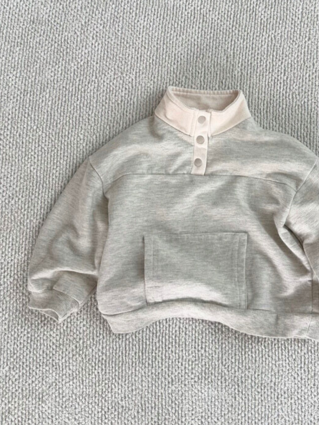 An EVERYDAY PULLOVER in heather grey with a funnel neck, buttoned placket, and front pocket, lying flat on a textured gray surface.