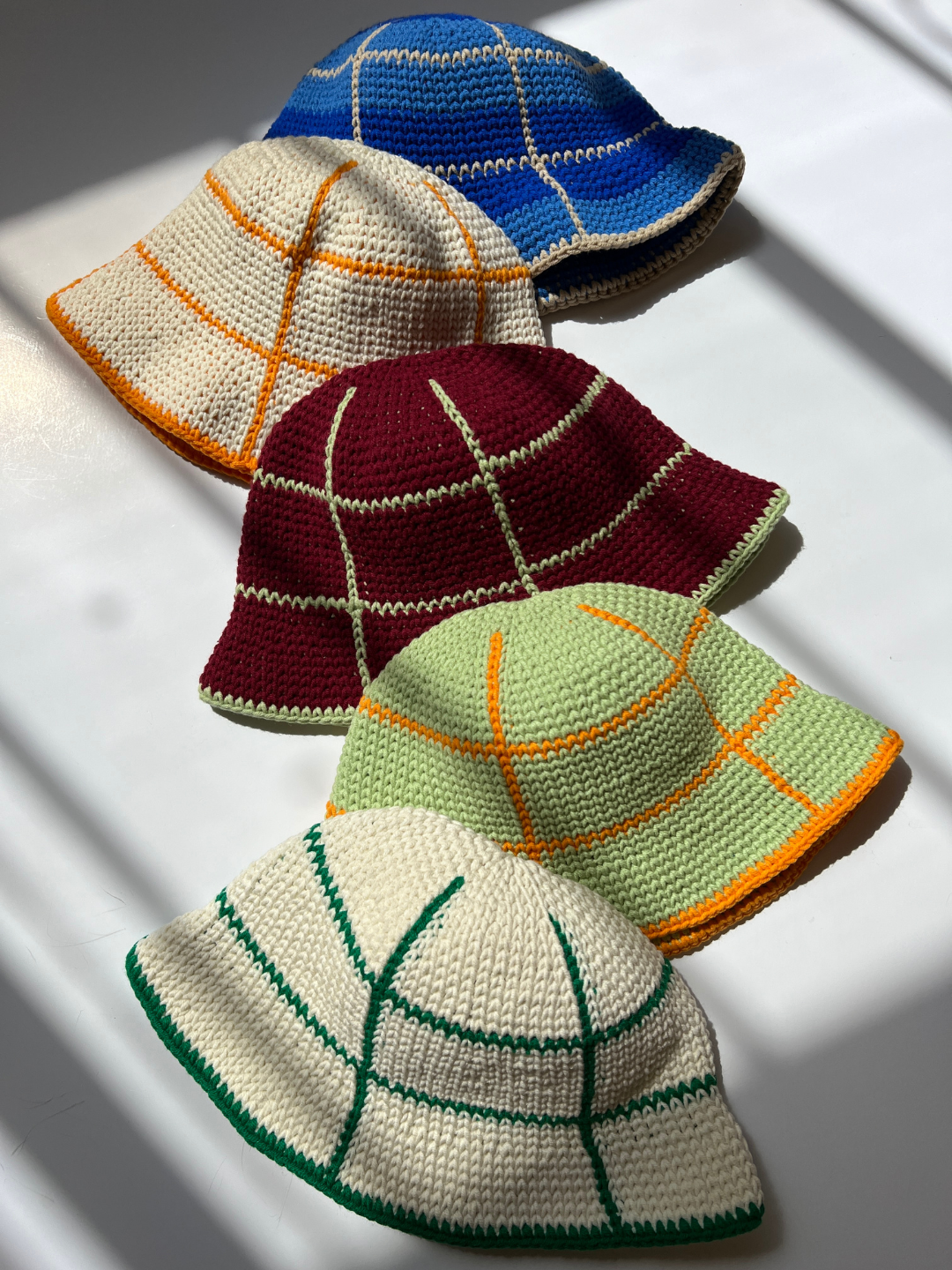 Five hand-crocheted GRID HATS - 3-6Y are displayed on a surface with sunlight casting shadows. The hats, made from cotton yarn, feature a grid design and come in different colors: blue with white and orange, white with orange and yellow, burgundy with white, green with orange, and white with green.
